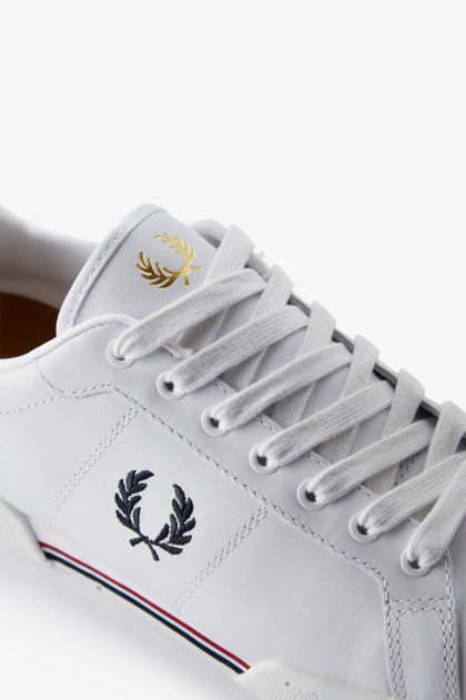 Close-up of a white sneaker featuring a gold and black laurel logo, with white laces and a striped detail on the sole.